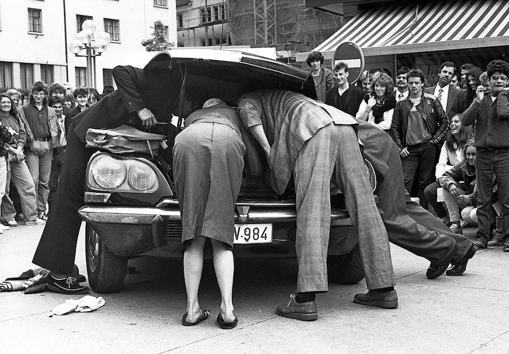 Schwarzweiss-Foto aus dem Jahr 1984, mit Theaterszene auf dem Zürcher Hechtplatz, drei Männer und eine Frau beugen sich über die geöffnete Motorhaube eines Autos, viel Publikum schaut zu und lacht.