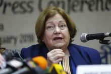 Zirka 70-jährige Frau mit blauem Blazer und rötlichen, halblangen Haaren sitzt vor mehreren Mikrofonen und spricht zu den Medien.